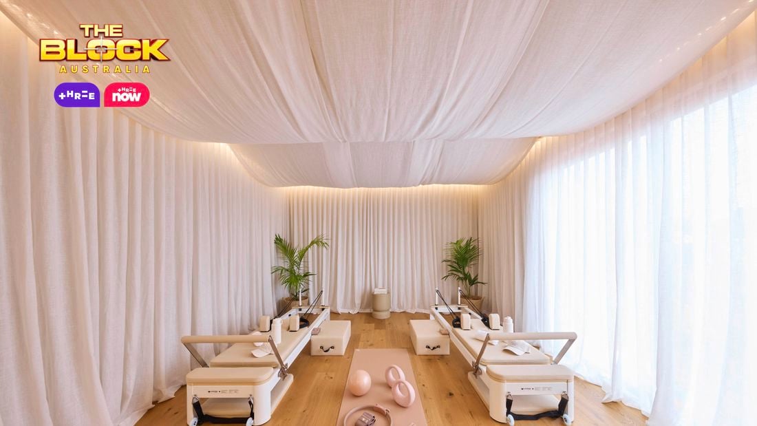 An elegantly designed Pilates studio with four reformer machines arranged symmetrically. The space features white draped curtains on the walls and ceiling, creating a serene ambiance. A pink exercise mat in the center holds pink fitness accessories including an exercise ball, resistance bands, and weights. Two potted plants sit at the far end, and the floor is light-colored wood. 