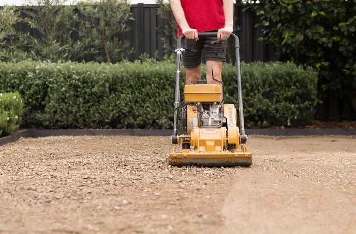 A person is using a compactor to level and compress the gravel base to create a firm and level foundation for the synthetic turf.