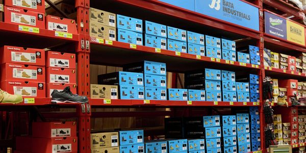 A workwear and safety footwear aisle in a large hardware warehouse, featuring red industrial shelving stocked with numerous boxed work boots and safety shoes. Blue Bata Industrials, Hard Yakka, Blundstone and DEWALT signage is displayed above the shelves, with products organised by brand and size.