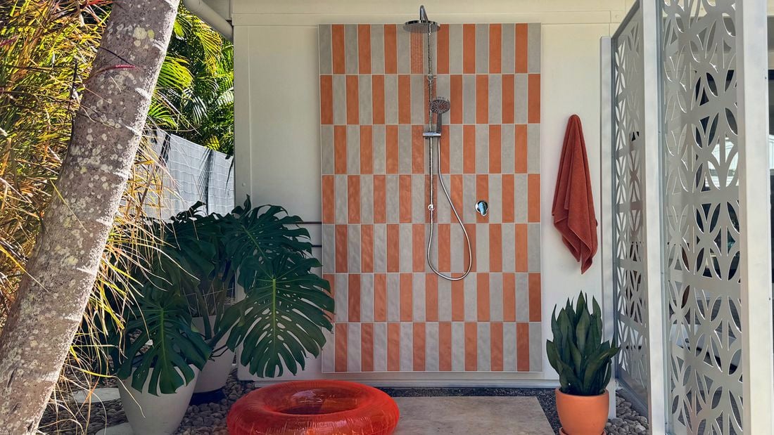 Outdoor shower with orange and white tile panel, overhead shower and hand shower. An orange towel, plants and pool toys sit nearby.