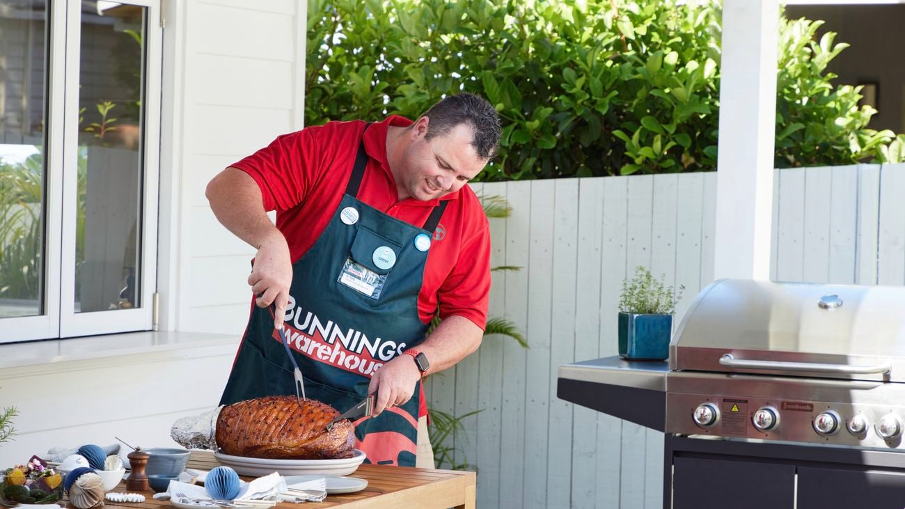 A Bunnings employee carves a Christmas ham on an outdoor table in front of a BBQ.