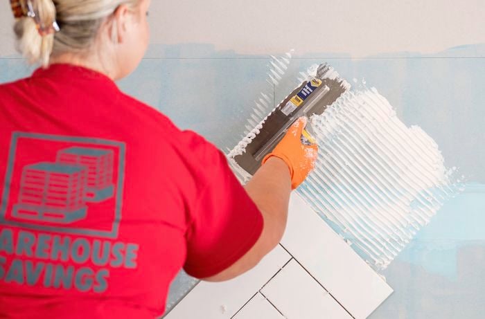 A Bunnings Team Member is spreading tile adhesive on a wall with a notched trowel above a row of herringbone tiles.