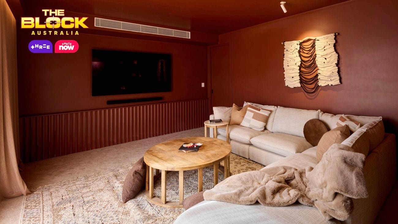 A warm-toned living room featuring a large beige sectional sofa with assorted pillows and throws. A round wooden coffee table sits in front of the sofa, facing a wall-mounted flat-screen TV. The walls are painted brown and decorated with an artistic wall hanging above the sofa. A patterned rug covers the floor.