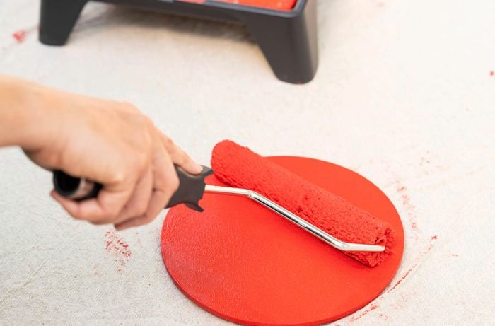 A person is applying bright red paint to a wooden placemat with a paint roller to create playful retro shapes for a DIY throwback mirror