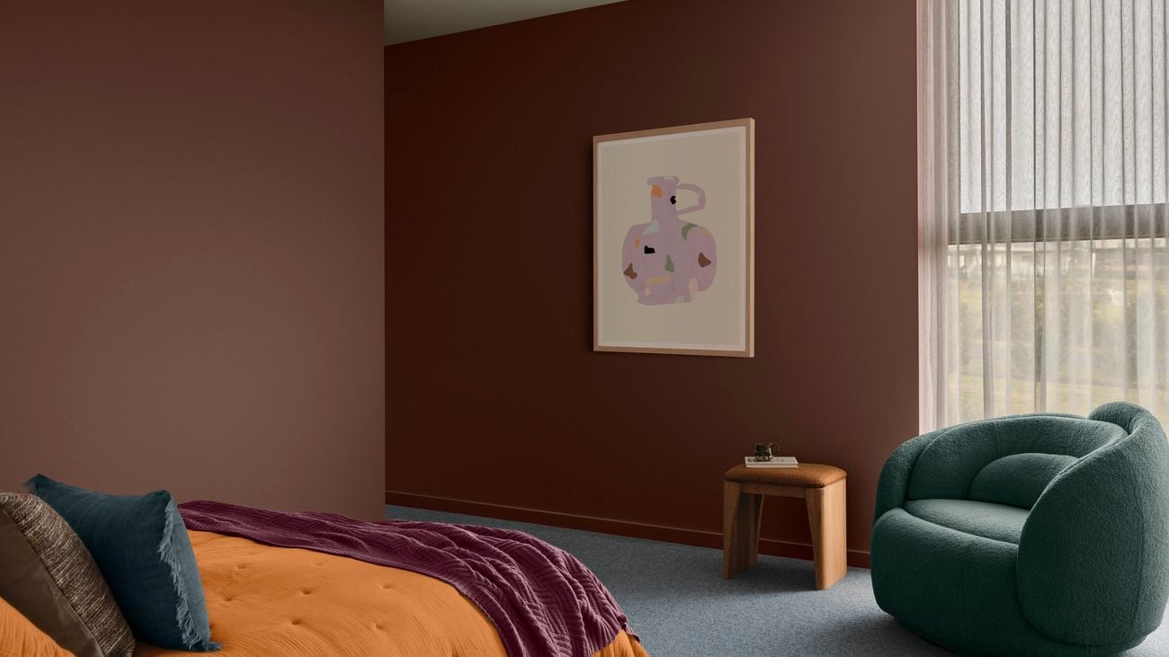 A stylised bedroom with Dulux Chocolate Treat wall paint, an orange bedspread, a teal chair and a painting of an abstract jug on the wall.