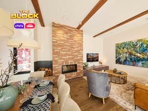 A modern living room featuring a central brick fireplace, exposed wooden ceiling beams, and a large abstract painting on the right wall. Furnished with a brown leather sofa, two gray armchairs, and a round wooden coffee table atop an area rug. A dining table set for a meal is visible to the left.