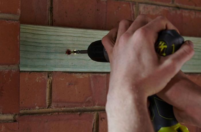 A person fixing a length of timber to a brick wall using a cordless drill