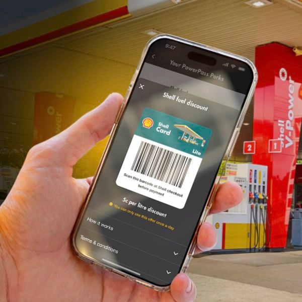 Hand holding a smartphone displaying a Shell fuel discount barcode in the PowerPass app, with a Shell service station visible in the background.