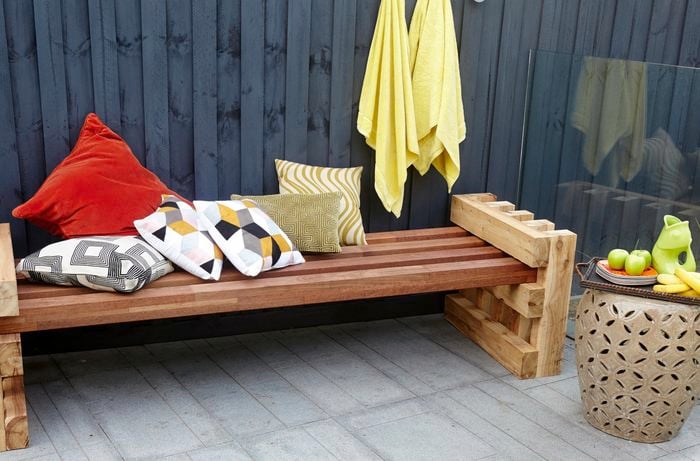 A completed outdoor day bed against a backyard fence with six throw cushions and a fruit tray nearby
