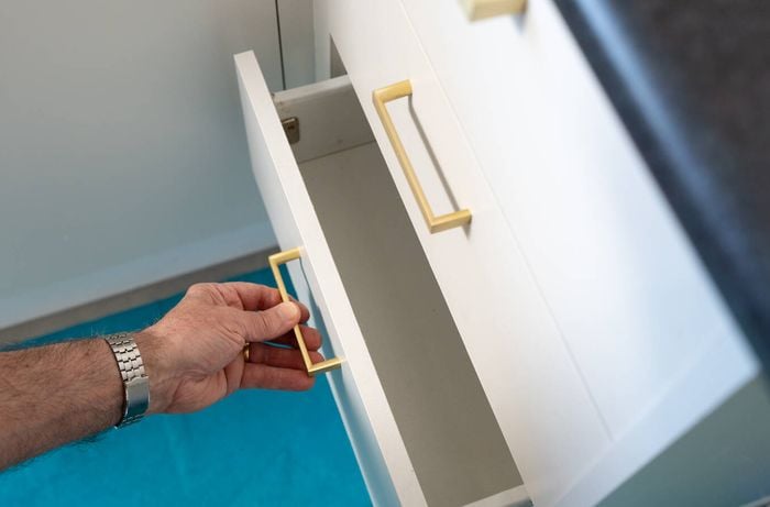 A person is opening a freshly painted white drawer with a gold handle in the kitchen.