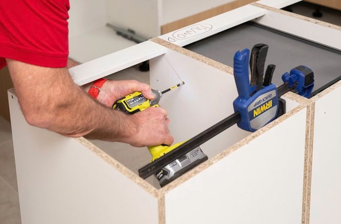 A person in a red t-shirt is using a drill to join the cabinets together during installation.