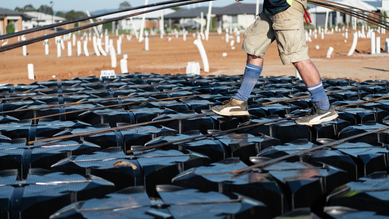 A tradie carrying a handful of rebar walks across the top of plastic foundation pods on a building site