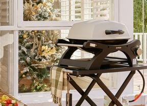A portable single‑burner gas barbecue on a foldable trolley with a gas bottle sits on a timber deck verandah beside a window; a decorated Christmas tree is visible inside, with outdoor furniture and trees in the background.
