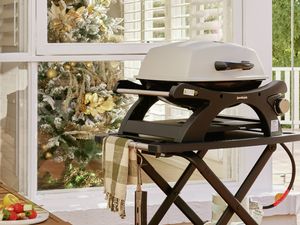 A portable single‑burner gas barbecue on a foldable trolley with a gas bottle sits on a timber deck verandah beside a window; a decorated Christmas tree is visible inside, with outdoor furniture and trees in the background.