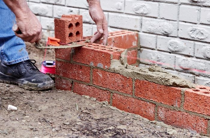 A person laying a third course of bricks parallel to a house wall