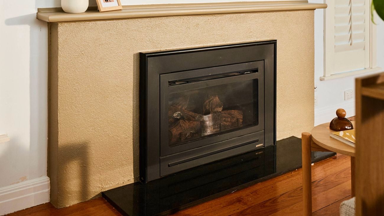 Modern fireplace with a black insert set into a beige surround, topped with a minimalist mantel holding vases and a framed photo, and a large abstract artwork hung above on a white wall.
