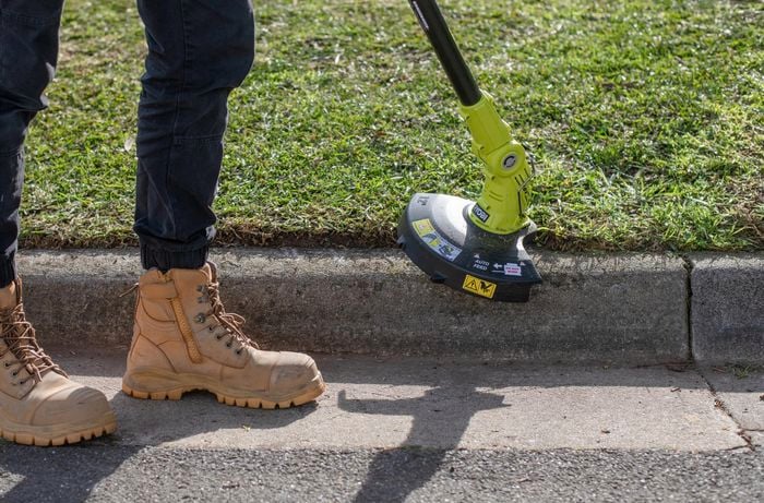 Person wearing work boots using whipper snipper tool on edge of lawn where it meets the path.