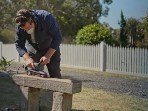 A man using a belt sander to sand back a heavy, hardwood timber sleeper seat outside