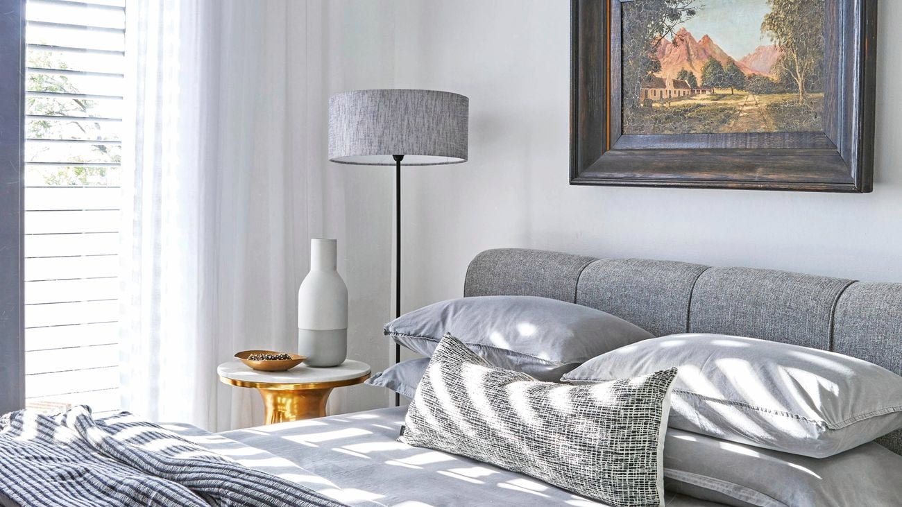 Bedroom with bed covered in a grey bedspread and assortment of throw rugs, with sunlight streaming through blinds on window.