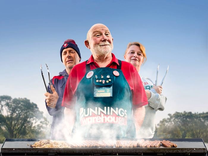 A Bunnings Team Member and 2 people in front of a BBQ with food on top of it.