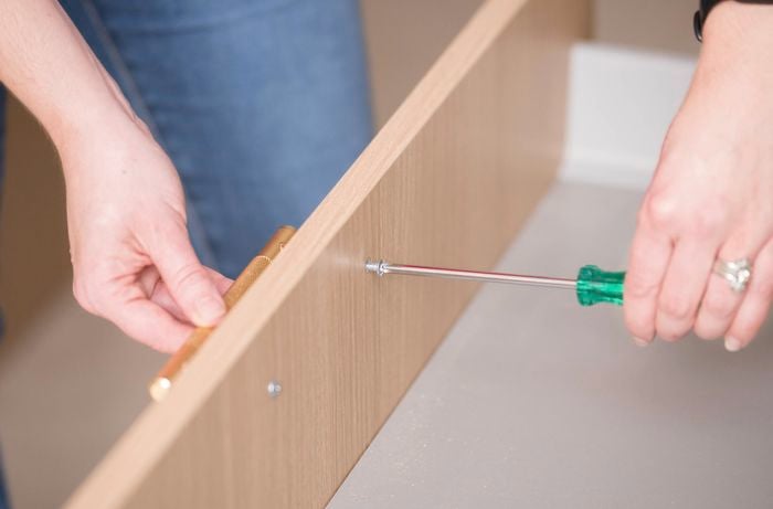 A person is using a screwdriver to attach a handle to a cabinet drawer to complete the kitchen.
