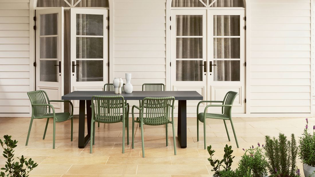 Outdoor dining table with olive green slatted chairs, on a paved patio in front of white French doors