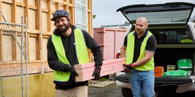 Workers wearing high-visibility vests carry timber planks from the back of a ute beside a partially built structure at a construction site.