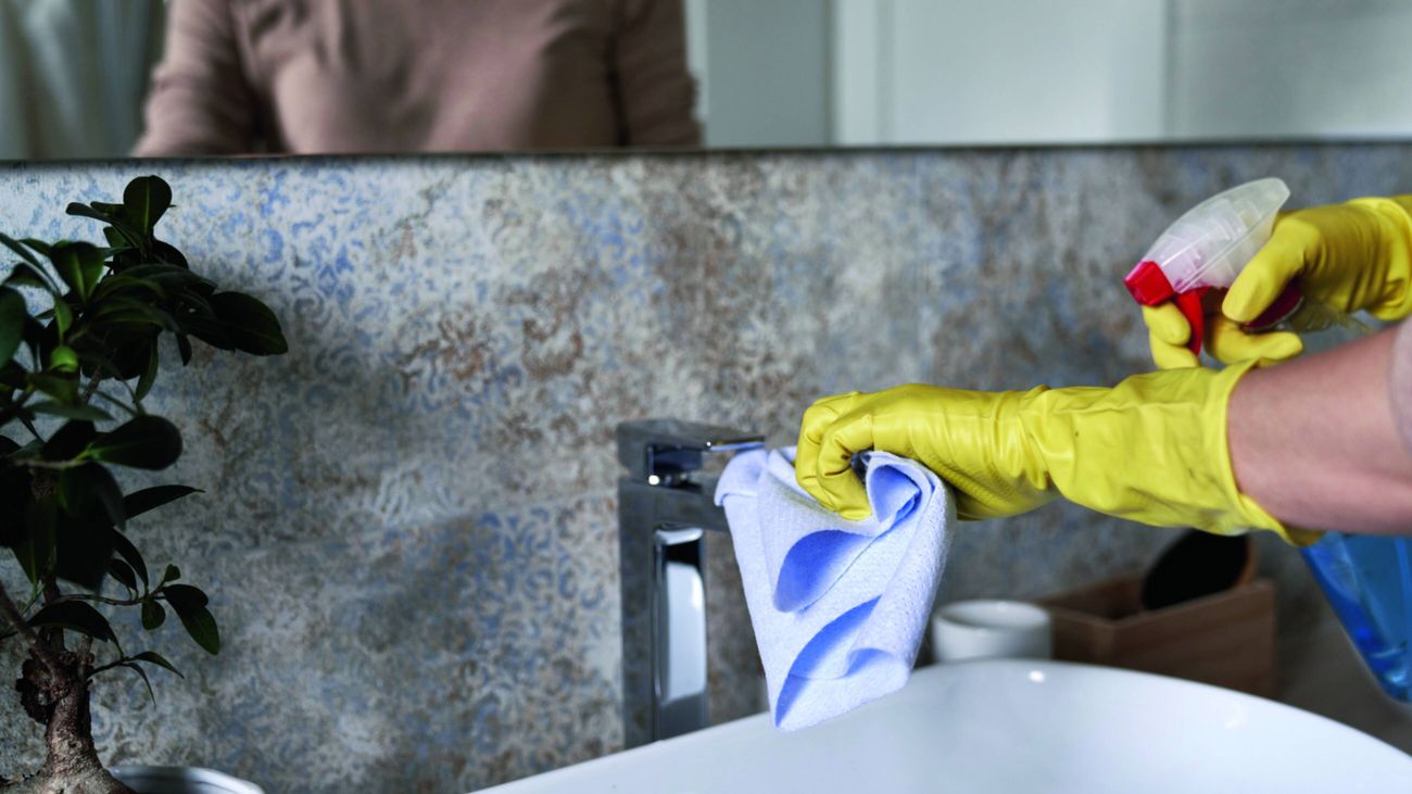 A person wearing yellow rubber gloves cleans a bathroom sink tap with a blue cloth and cleaning spray, with a mirror reflecting them in the background.