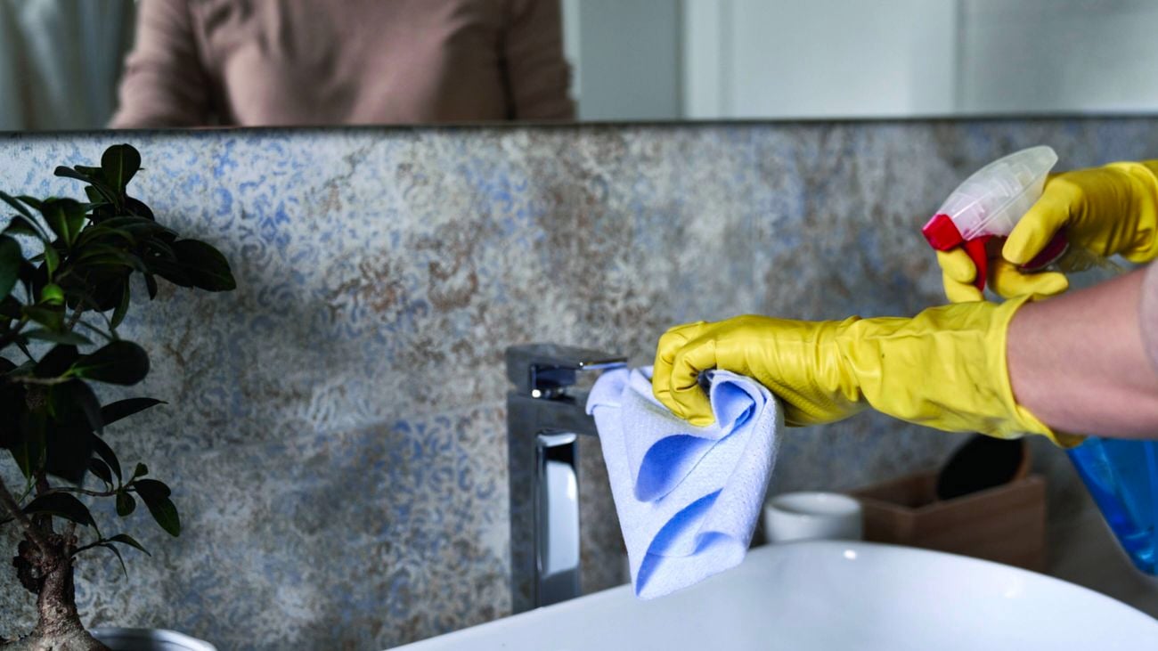 A person wearing yellow rubber gloves cleans a bathroom sink tap with a blue cloth and cleaning spray, with a mirror reflecting them in the background.