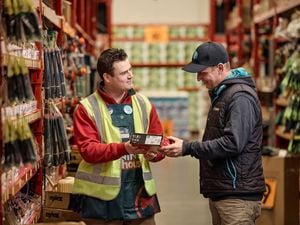 Bunnings team member handing a boxed product to a customer in a hardware store aisle with tools displayed on shelving.