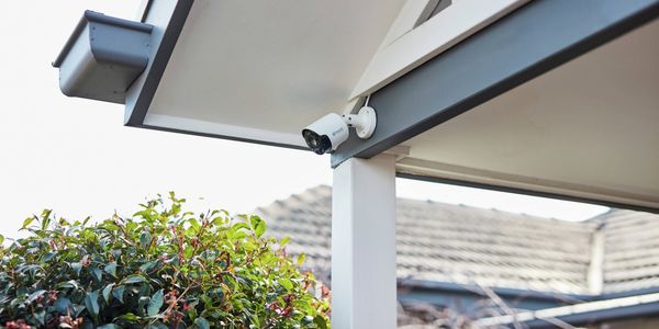 White security camera installed under a house roof eave above an outdoor area, with shrubs and neighbouring rooftops visible in the background.