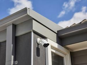 Close-up of a house corner with a white cylindrical security camera mounted near the roofline, grey siding and trim, and a partially visible window below against a blue sky with clouds.