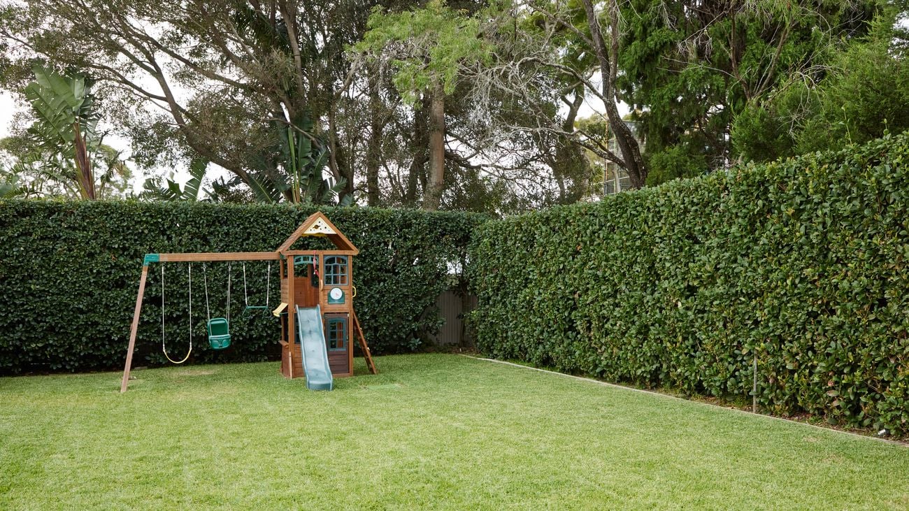 A wide-angle shot of a backyard with well-kept grass, a wooden playhouse with slide and trimmed, hedges surrounding it