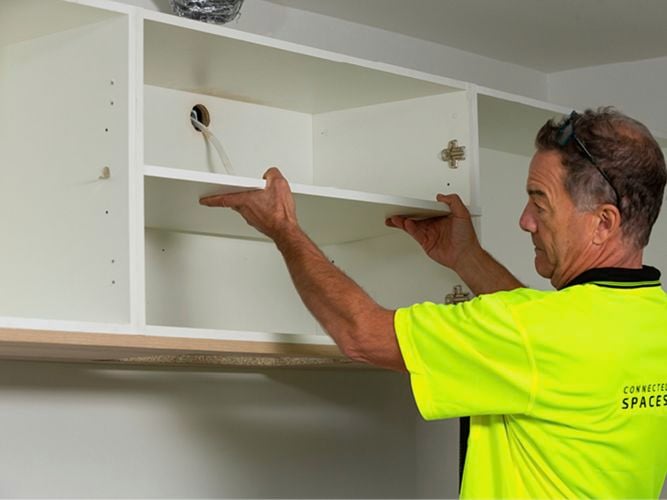 work man in green high vis top putting in kitchen overhead shelving