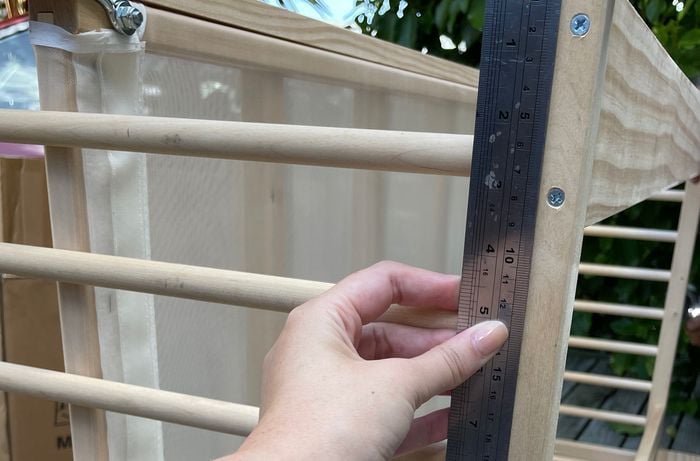 Hand holding a metal ruler next to the frame of the wooden cot