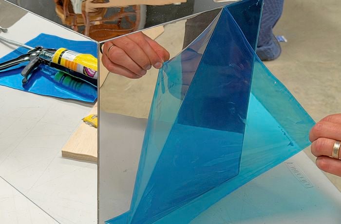 A hand peels a blue protective film from a mirrored surface, a caulking gun is visible in the background.