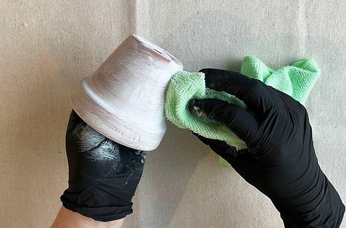  A pair of hands wearing black gloves holds a painted terracotta pot and wipes off a whitewash using a green cloth, on a drop sheet