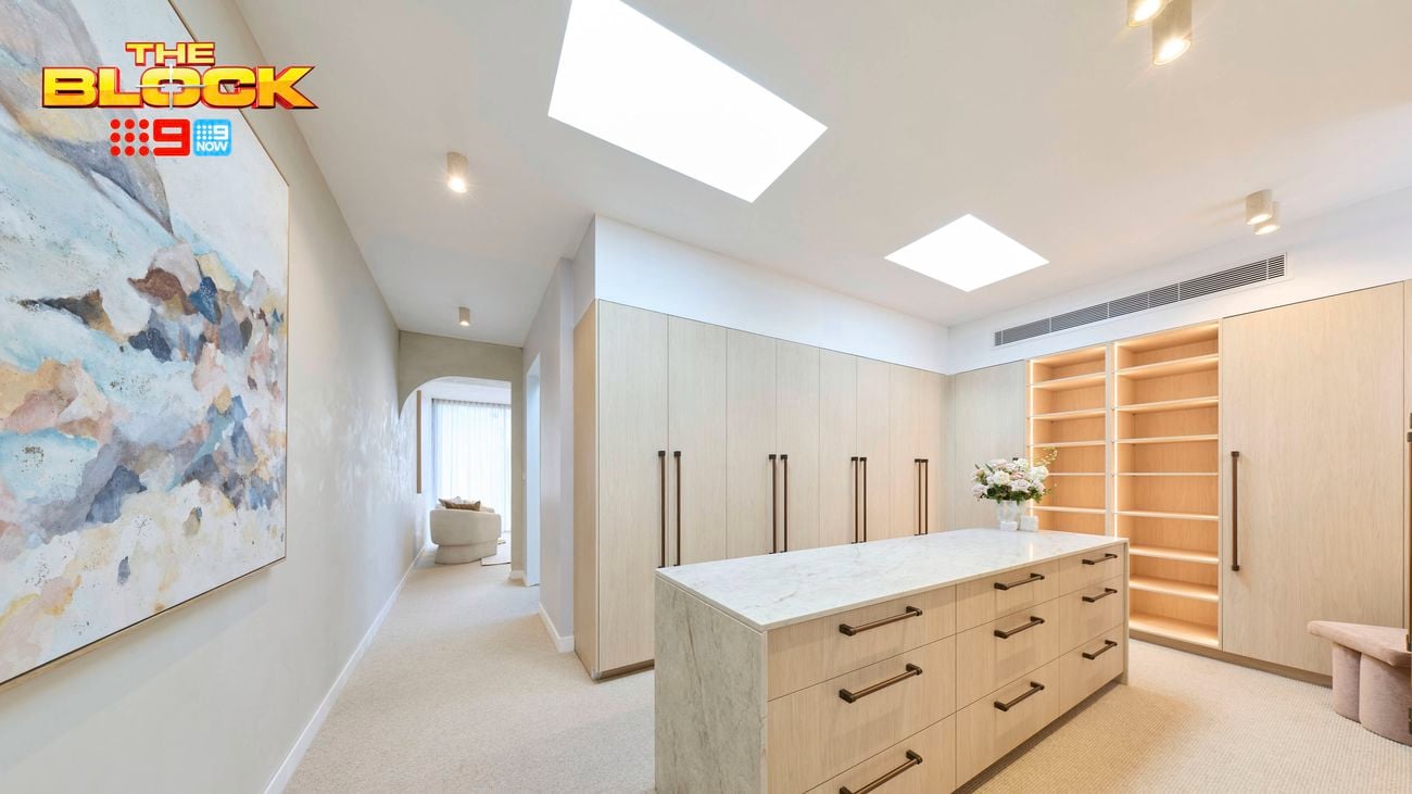 Spacious walk-in wardrobe with light timber cabinetry, central island drawers, open shelving, skylights and a large abstract artwork.