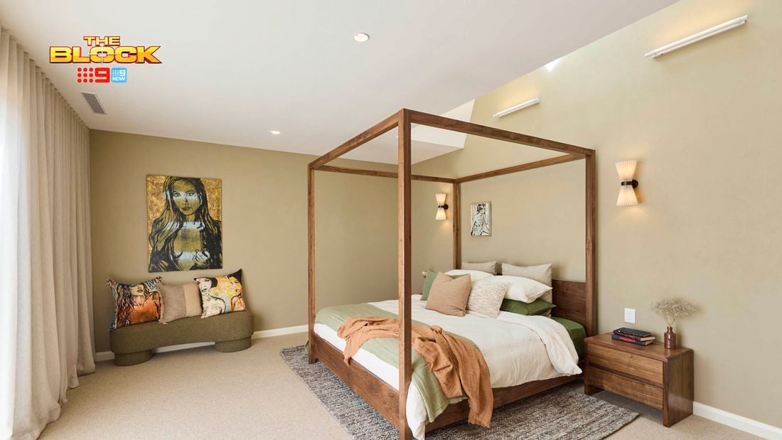 A stylish bedroom featuring a central four-poster bed with white and green bedding, multiple pillows, and a brown throw blanket. A wooden nightstand with books and decor sits to the right, while a cushioned bench with decorative pillows lines the left wall. The room includes a textured rug, light beige walls, wall-mounted lights, and artwork above the bench and bed. Sheer curtains cover large windows, allowing natural light in. 