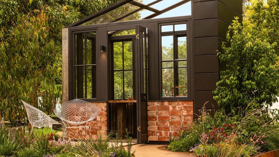 Modern black-framed greenhouse garden room with brick base, surrounded by lush plantings.