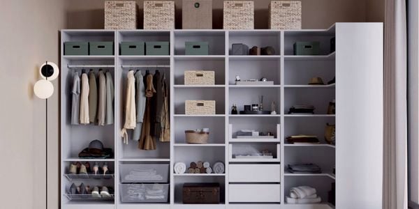 A large open wardrobe unit with shelves and hanging rails holding clothes, shoes, folded towels, storage boxes, wicker baskets, drawers and boxes on top. A floor lamp stands on the left.