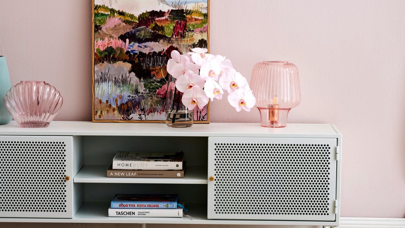 Wooden coffee table in foreground, cabinet in background with two pink, glass decorations, pink flowers and a painting on top.