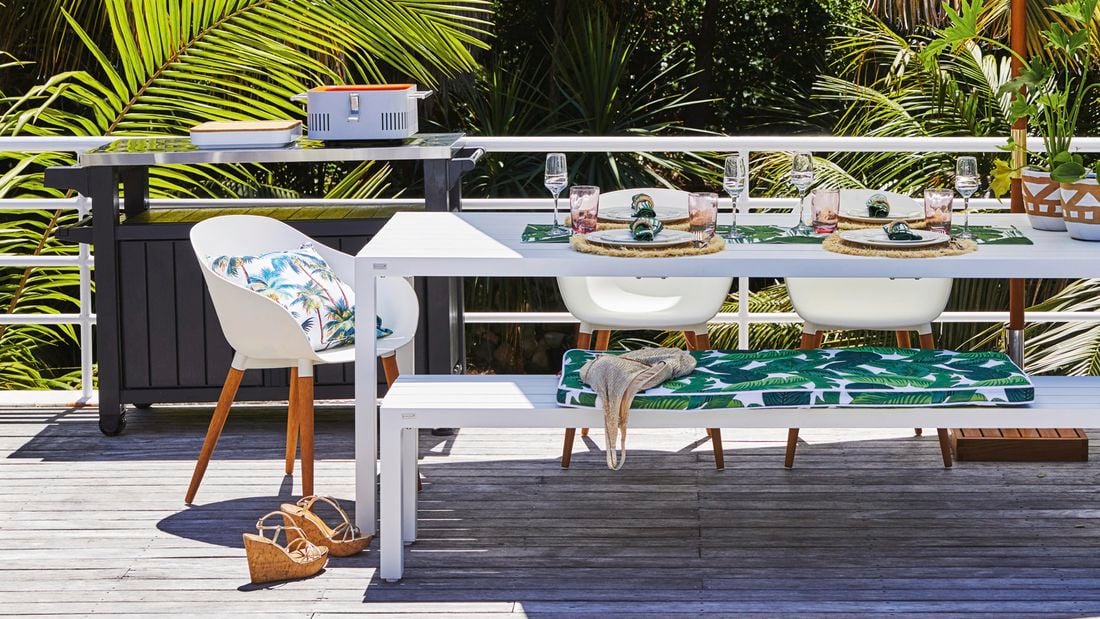 Miami inspired Outdoor living area in a tropical-like area with a long white table fully set, accompanied by matching bench seats. In the background, at the edge of the outdoor deck is a portable table with food dishes on it.