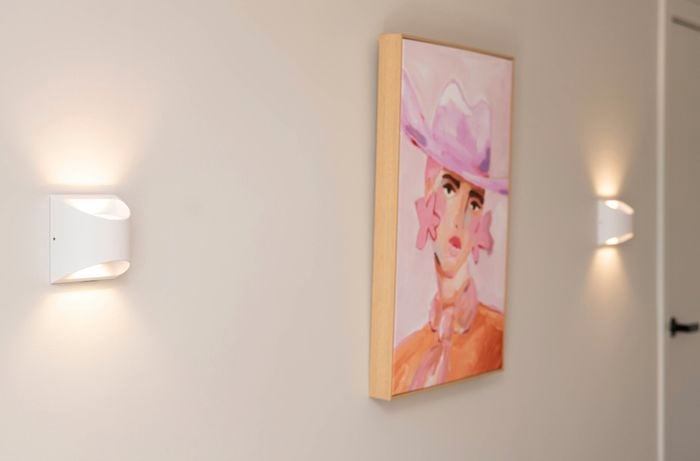 The image shows a hallway with soft wall lighting and a vibrant pink-themed portrait of a woman wearing a hat.