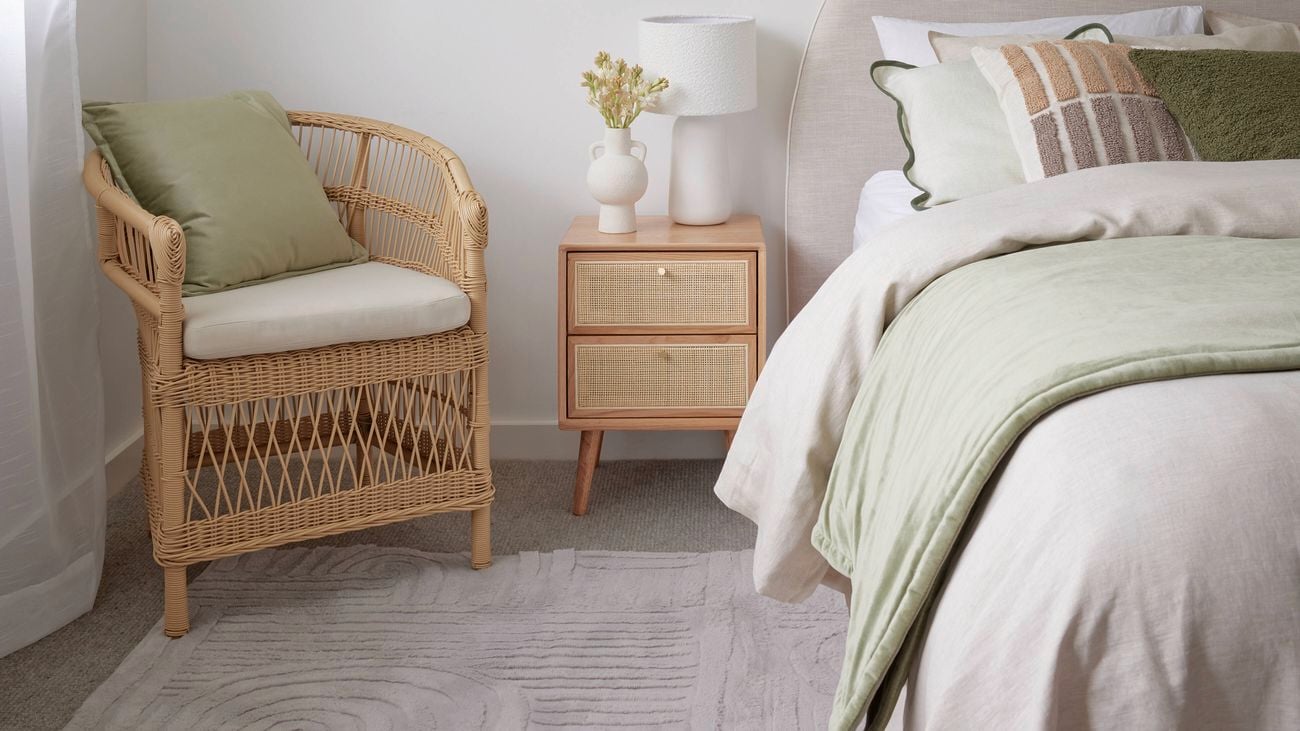 Bedroom with woven chair, bed with beige and green linen and pillows, a brown bedside table with a white lamp and a grey patterned rug on the floor.
