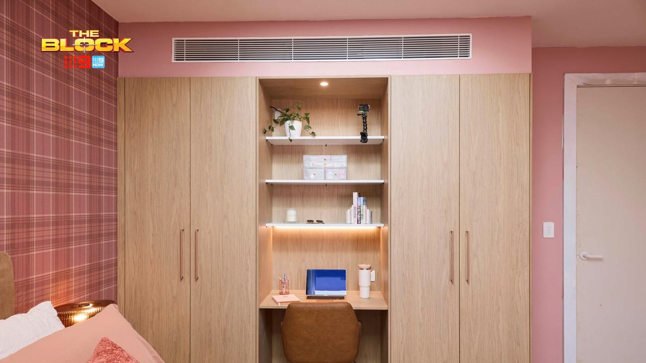 Pink bedroom with a check wallpapered wall and a double bed with pink linens. Opposite is a built in wardrobe with a desk in the middle.