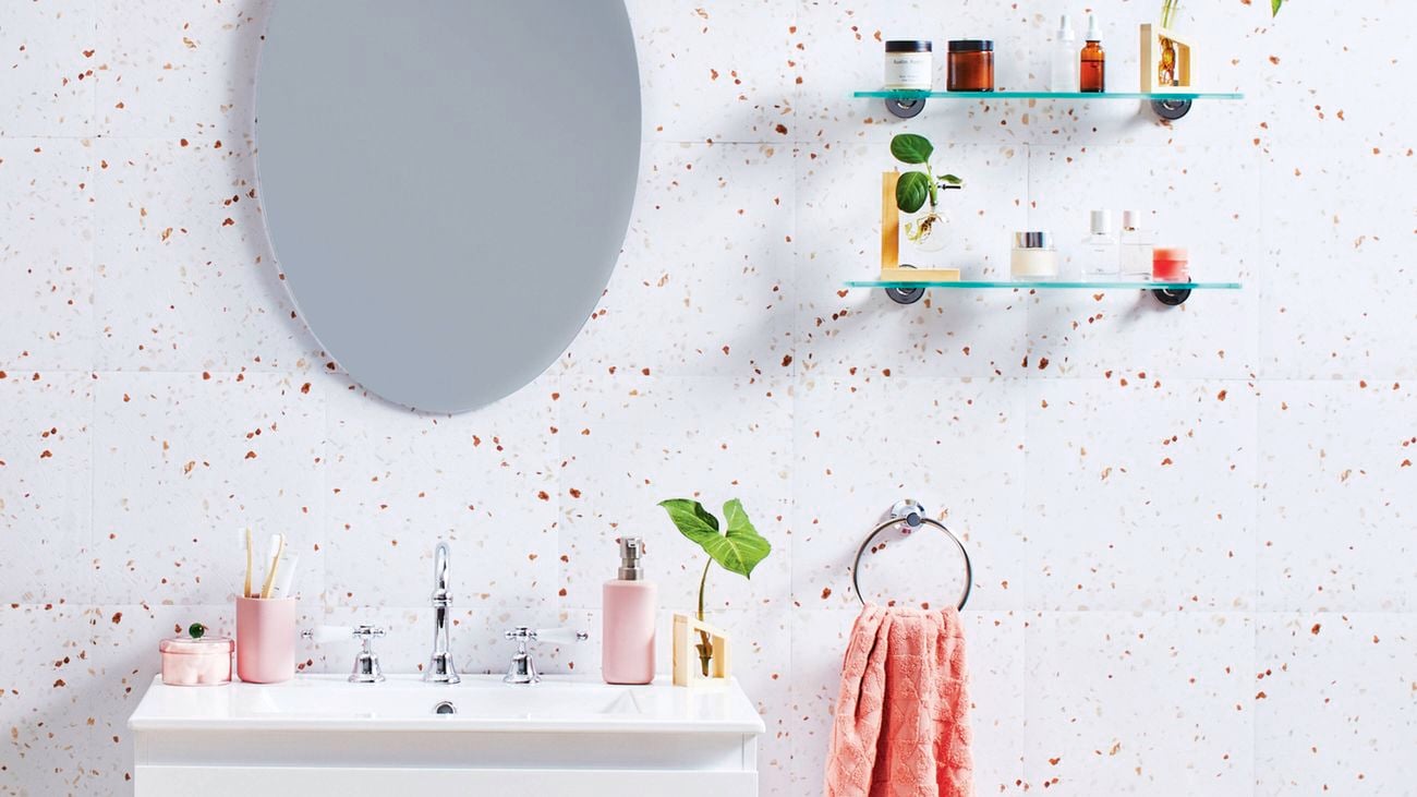 A light-coloured bathroom with speckled tiling, a basin with light pink accessories, a salmon coloured hand towel, an oval mirror and two floating glass shelves in a decluttered bathroom.