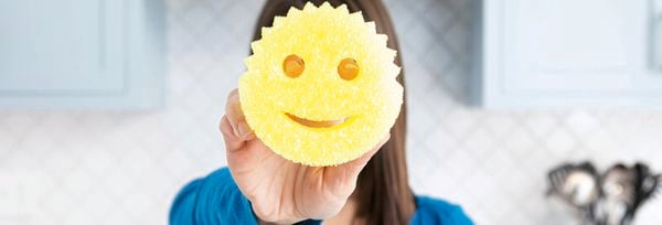 A woman holding a yellow Scrub Daddy sponge in front of her face, with a blue kitchen in the background.