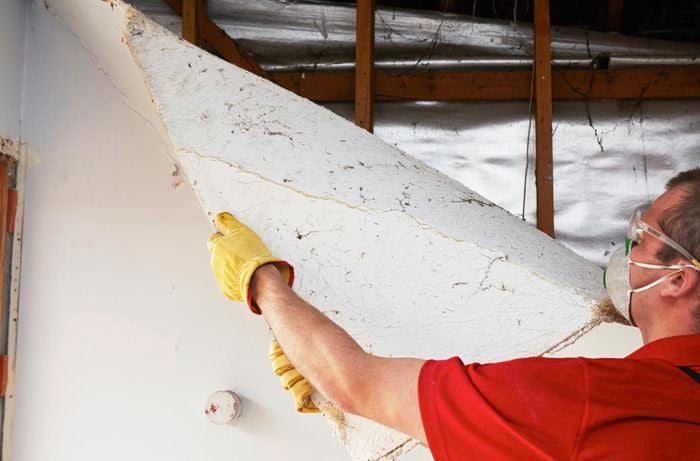 A person wearing protective gear pulling a sheet of plaster off a stud wall