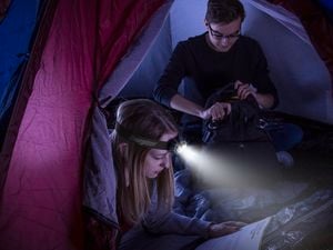  A male and a female sitting inside a tent at night. The woman is using a head torch to shine a light on the book in her hand and the man is looking through his backpack